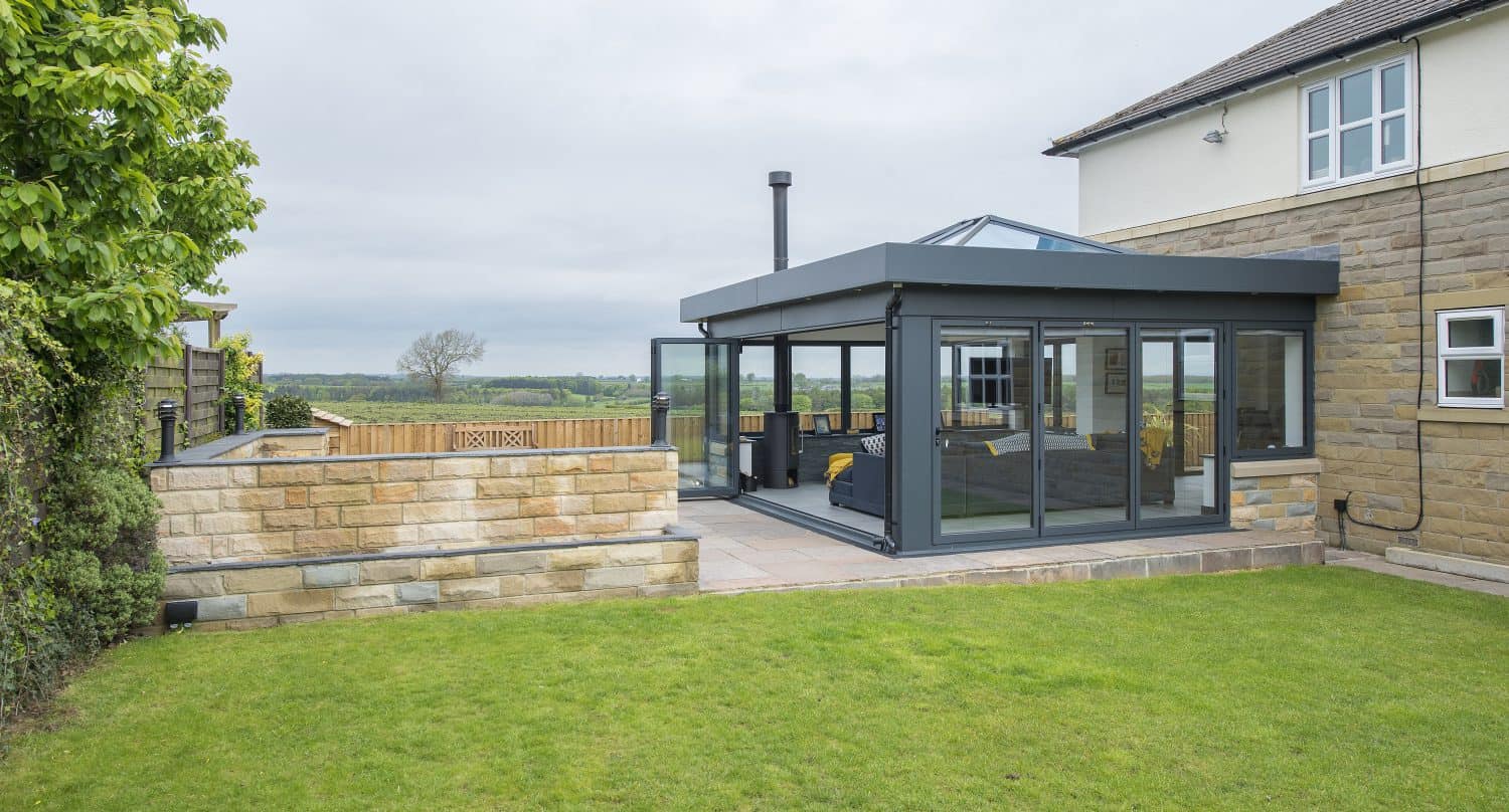 A modern glass conservatory with a black frame and bifold doors is attached to a stone and white house, overlooking a green lawn and countryside. The patio area features outdoor seating and a low stone wall.