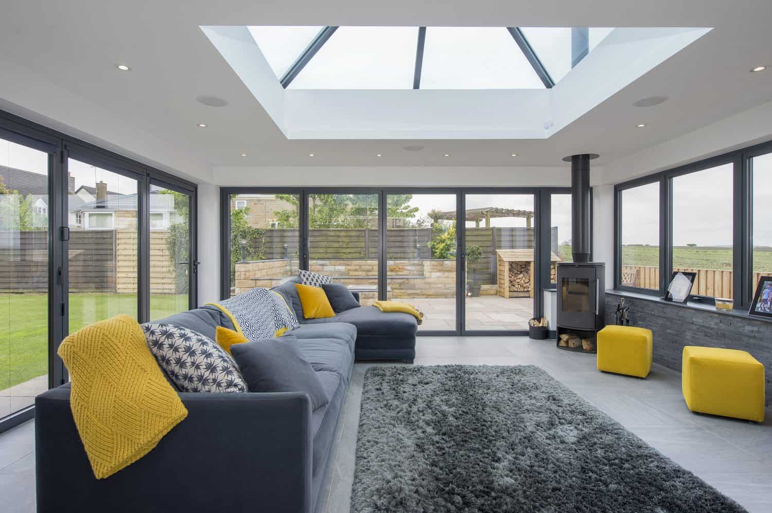 Modern sunroom with large glass roofing, grey sofas accented with yellow throws and cushions, grey rug, yellow stools, and floor-to-ceiling bifold doors opening to a patio and garden.