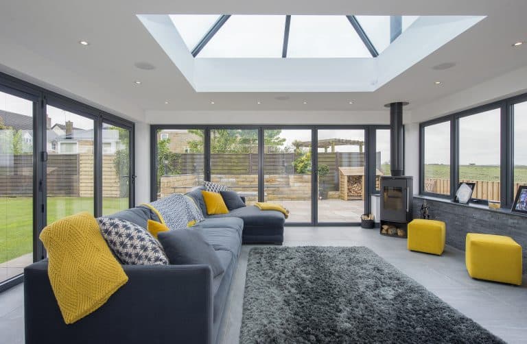 Modern sunroom with large glass roofing, grey sofas accented with yellow throws and cushions, grey rug, yellow stools, and floor-to-ceiling bifold doors opening to a patio and garden.