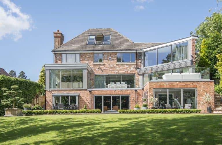 Modern three-story brick house with large glass windows and bespoke glass solutions, featuring balconies and a well-manicured lawn surrounded by greenery under a clear blue sky.
