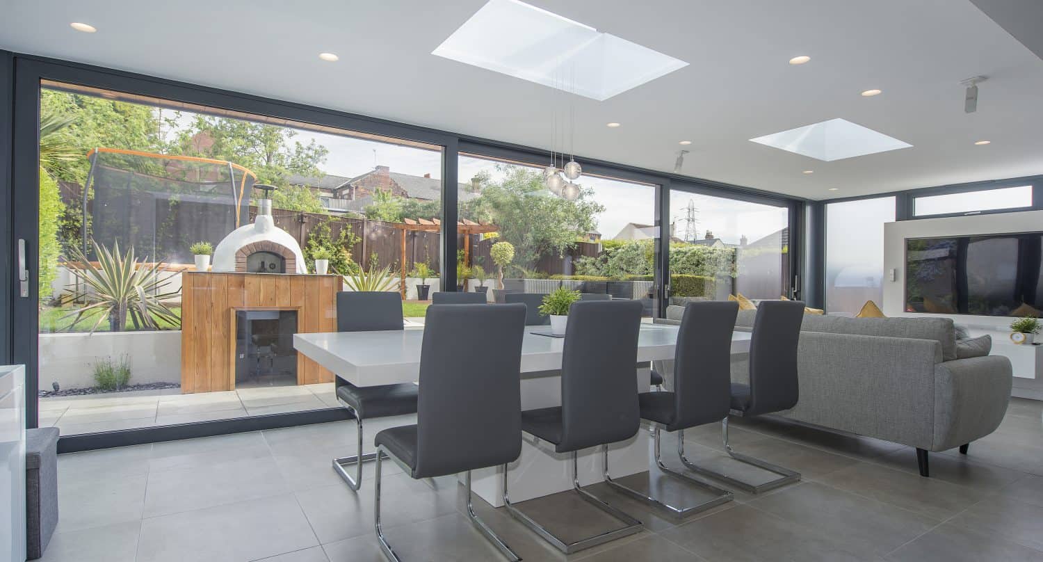 Modern open-plan dining and living area with large glass bifold doors overlooking a garden patio, pizza oven, and greenery. Contemporary furniture, skylights, and neutral tones create a bright, airy atmosphere.