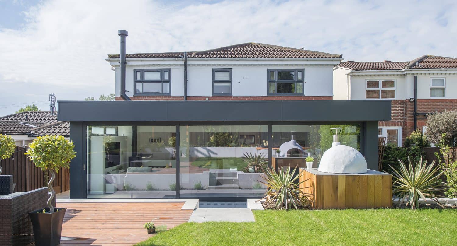 A modern house with bespoke glass solutions, a large glass extension, flat roof, and a spacious backyard featuring green grass, a wooden deck, potted plants, and a built-in outdoor pizza oven.