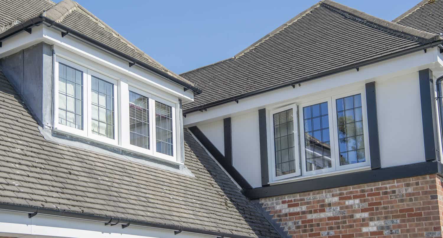 Close-up view of a modern house with gabled roofs, gray shingles, brick and white exterior walls, and large multi-pane windows featuring bespoke glass solutions under a clear blue sky.