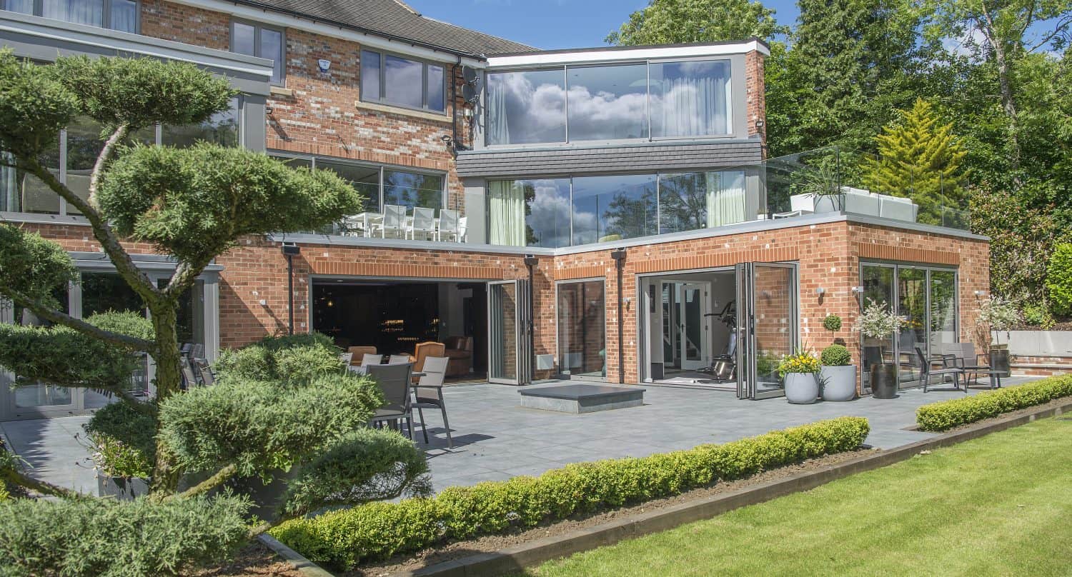 Modern brick house with large glass windows and front doors, a spacious patio with outdoor seating, potted plants, manicured lawn, and sculpted trees, set against a backdrop of green trees and blue sky.
