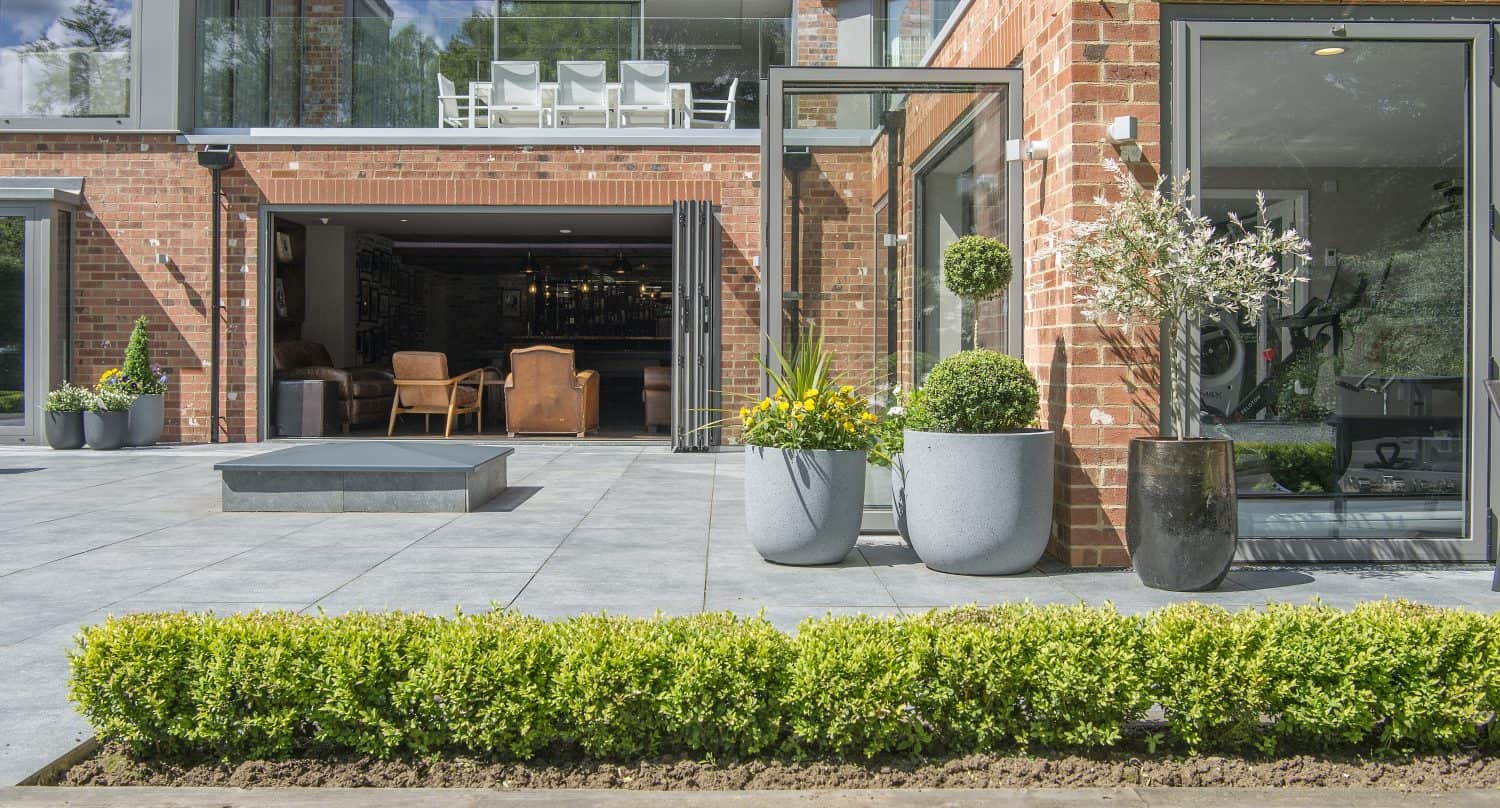 A modern brick house with bifold doors opens to a spacious patio, potted plants, and neatly trimmed hedges in the garden. Indoor seating and a home gym are visible through the open doors.