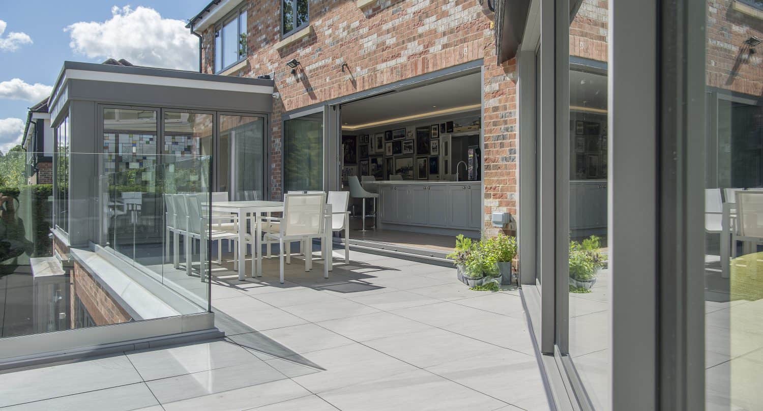 Modern patio with large tiled flooring, white outdoor dining set, and bifold doors opening to a spacious kitchen in a red-brick house, on a sunny day with blue sky and clouds.