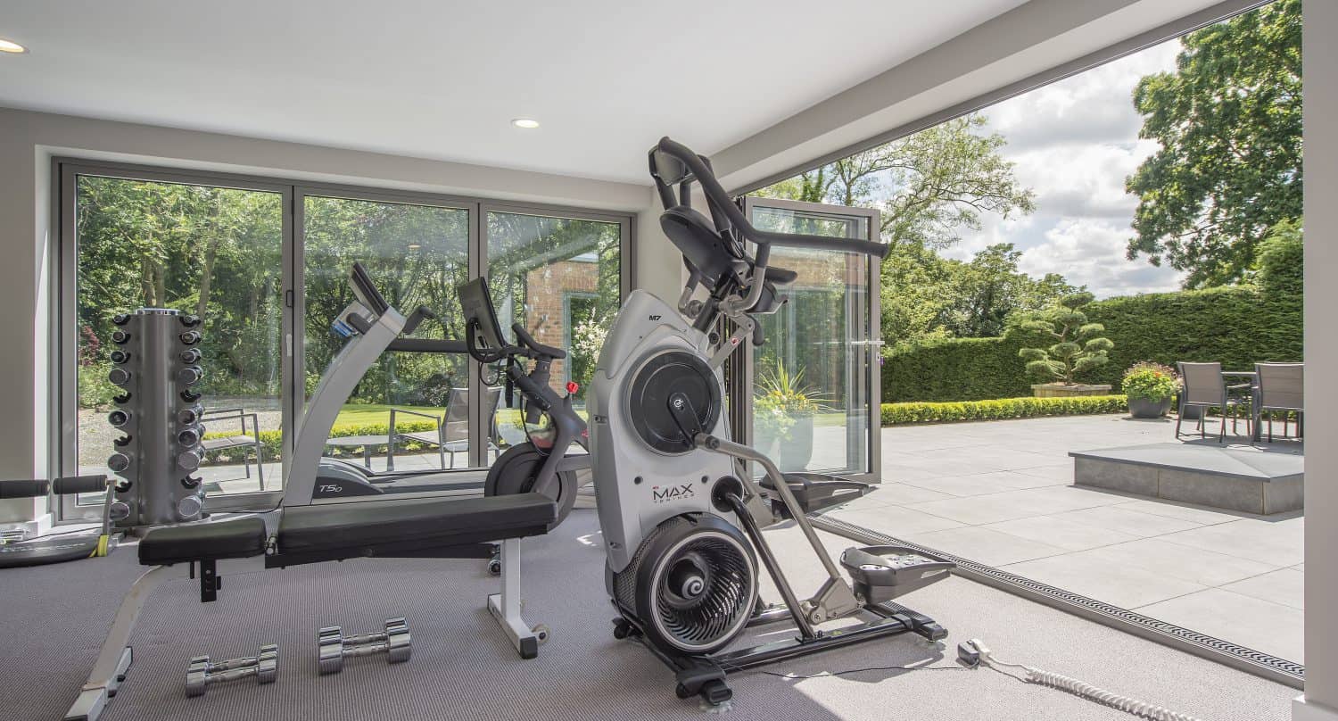 Home gym with exercise equipment including a stationary bike, elliptical machine, and dumbbells by large windows and bespoke glass solutions, overlooking a patio and garden with outdoor furniture and greenery.