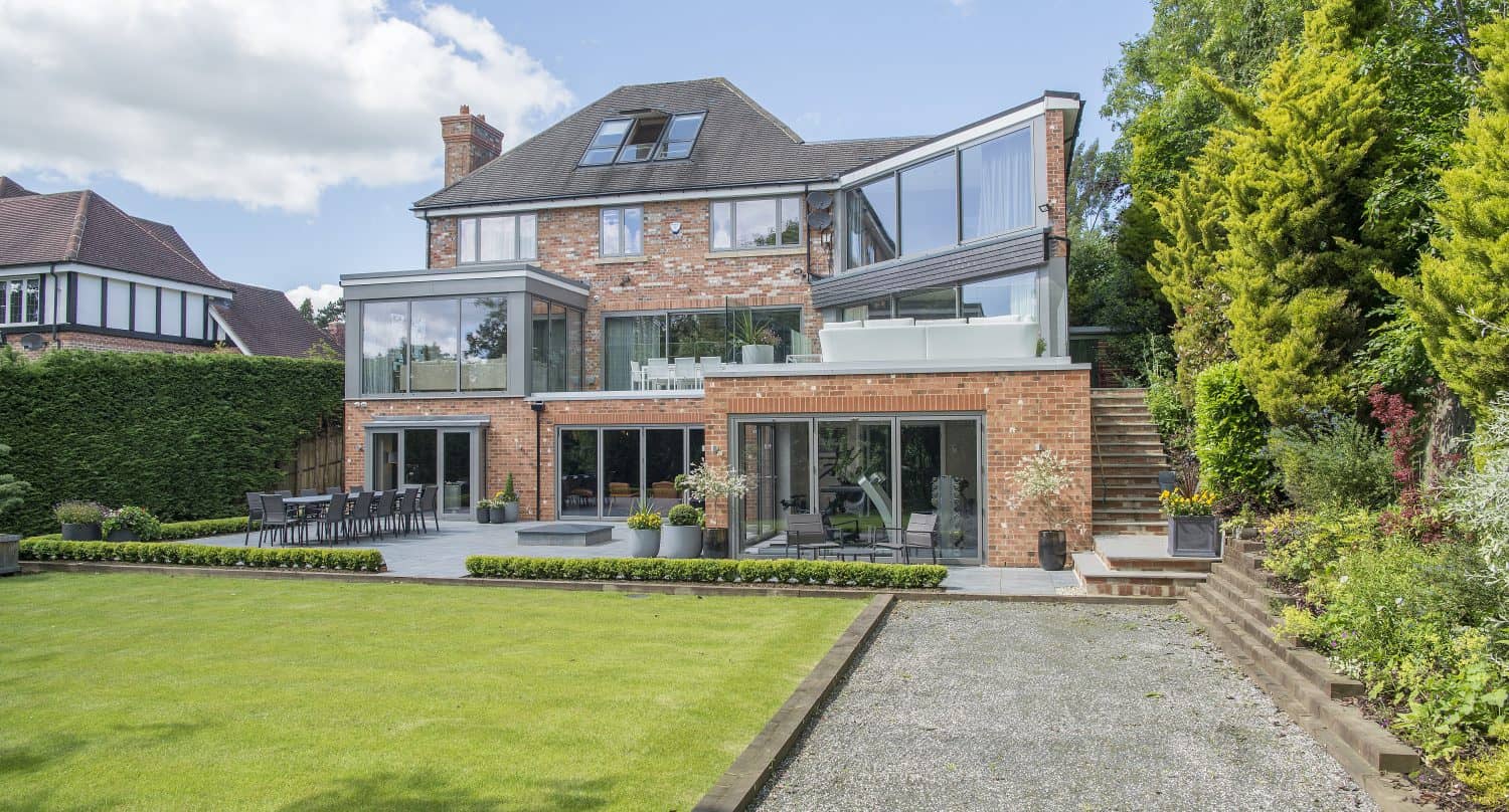 A large modern brick house with multiple glass balconies, bifold doors, and patios overlooking a well-kept lawn and garden, surrounded by hedges and trees under a partly cloudy sky.