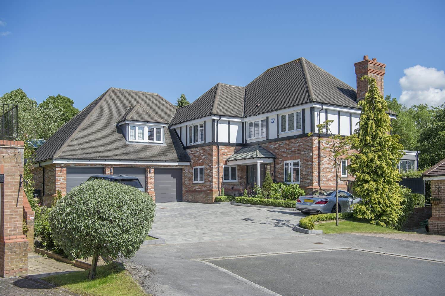 A large two-story house with brick and white exterior, black trim, gray roof, bespoke glass solutions including sliding doors, attached garage, and paved driveway with two parked cars, surrounded by greenery under a clear blue sky.