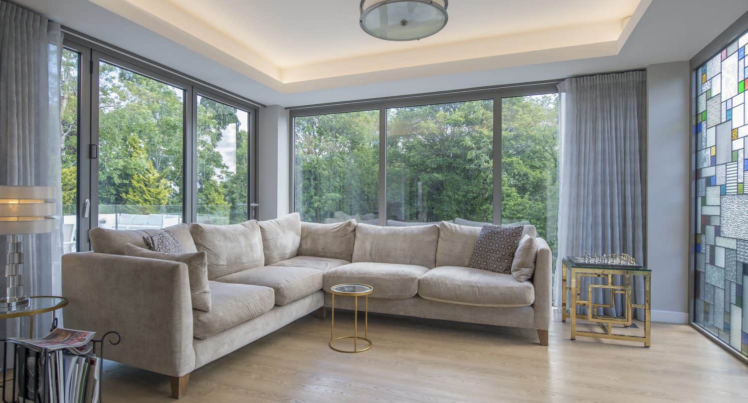 Modern living room with large windows, a beige sectional sofa, light wood floor, glass side tables, and a stained glass accent wall. Sliding doors open to lush greenery outside, filling the softly lit space with natural beauty.