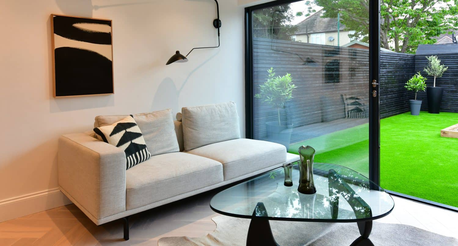 Modern living room with a light gray sofa, black-and-white pillows, abstract wall art, and a glass coffee table on a light rug. Large bespoke glass sliding doors open to a green backyard with plants and outdoor seating.