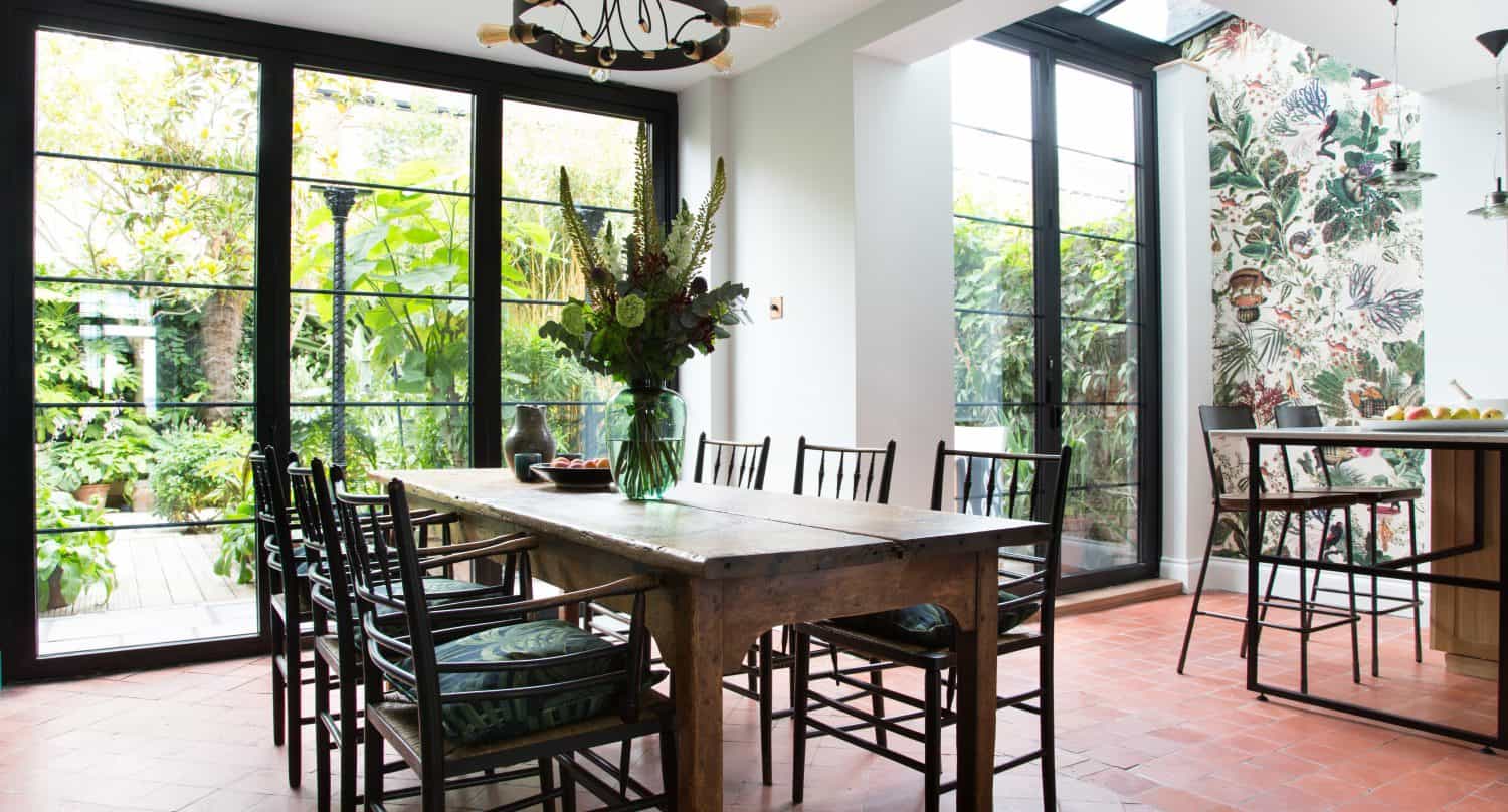 Bright dining area with large windows, wooden table and black chairs, bifold doors opening to green plants outside, floral wallpaper on one wall, and a vase of flowers as the centerpiece on the table.