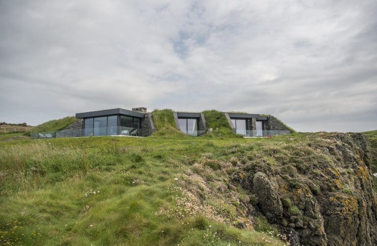 A modern house with large glass windows and sleek bifold doors is built into a grassy hillside, blending with the landscape. The building overlooks rocky cliffs and is surrounded by lush green grass under a cloudy sky.