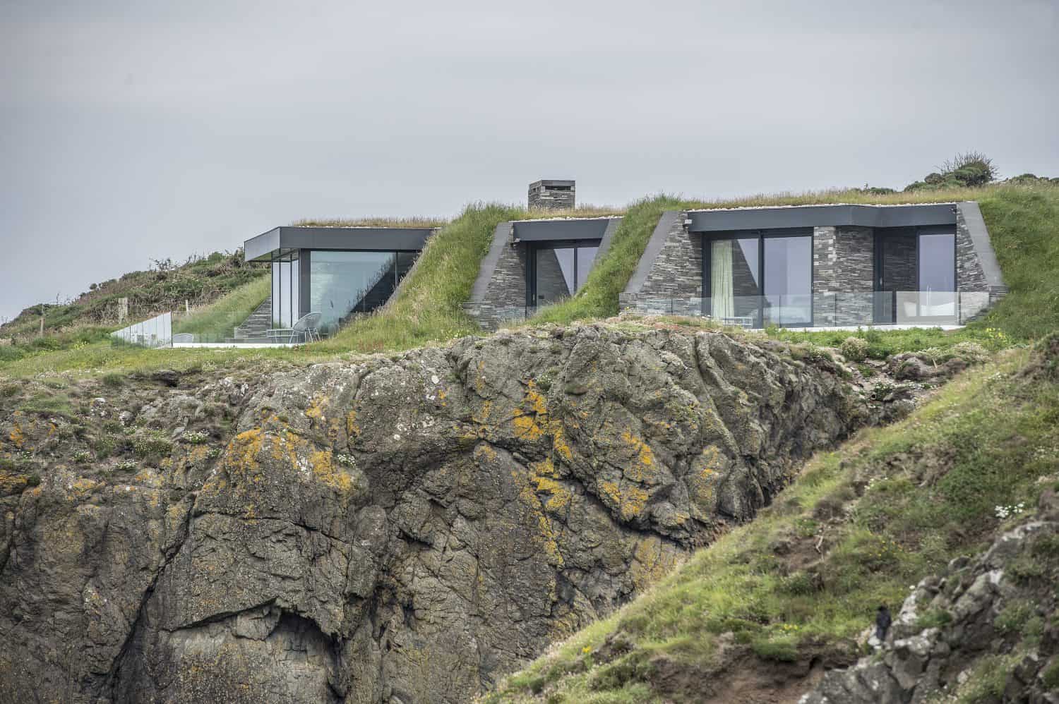 Modern houses with large glass windows and sliding doors are built into a grassy hillside, overlooking a rugged cliff edge with patches of moss and vegetation, under an overcast sky.