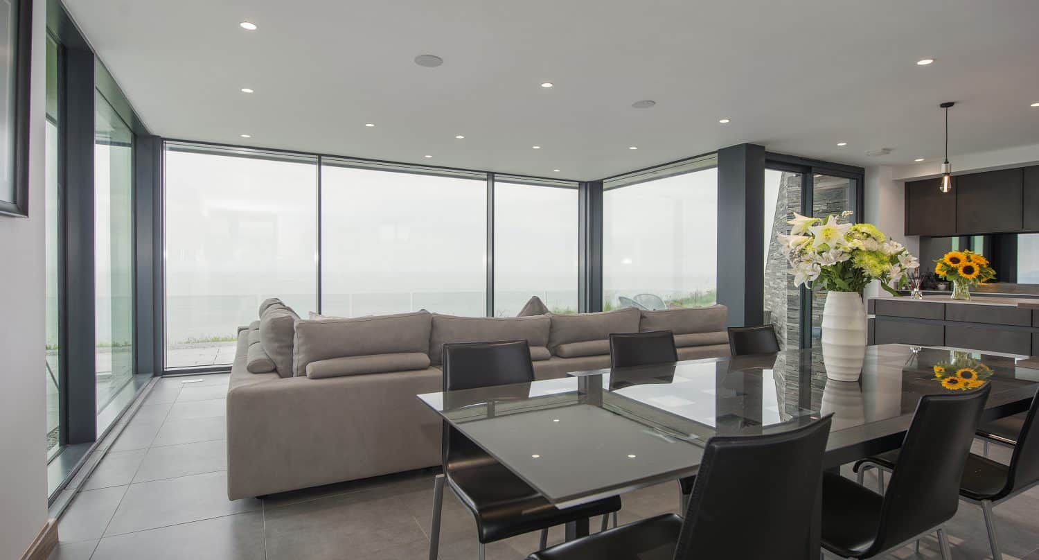 Modern open-plan living and dining area with glass walls and sliding doors offering an ocean view, a beige sectional sofa, glass dining table with black chairs, and vases of flowers on the table and kitchen island.