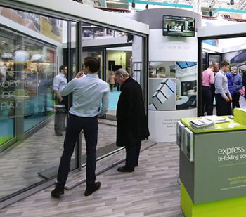 People at an indoor exhibition observe and interact with large sliding doors. Booths display information and samples, including innovative front doors, while a TV screen shows related visuals in a modern, professional setting.