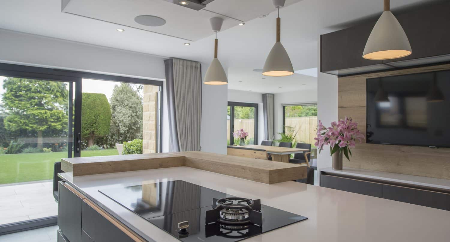 Modern kitchen with a sleek black gas stove on a white island, pendant lights overhead, large windows and bespoke glass solutions like sliding doors leading to a green garden, and a bouquet of pink flowers near a wall-mounted TV.