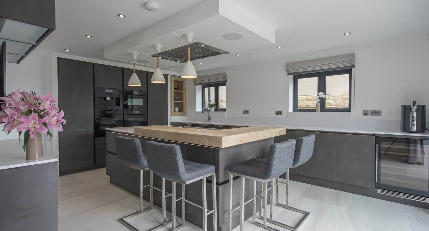 Modern kitchen with a large island, four gray bar stools, built-in appliances, pendant lights, dark cabinets, and light countertops. Bespoke glass solutions like bifold doors and two windows fill the space with natural light and style.