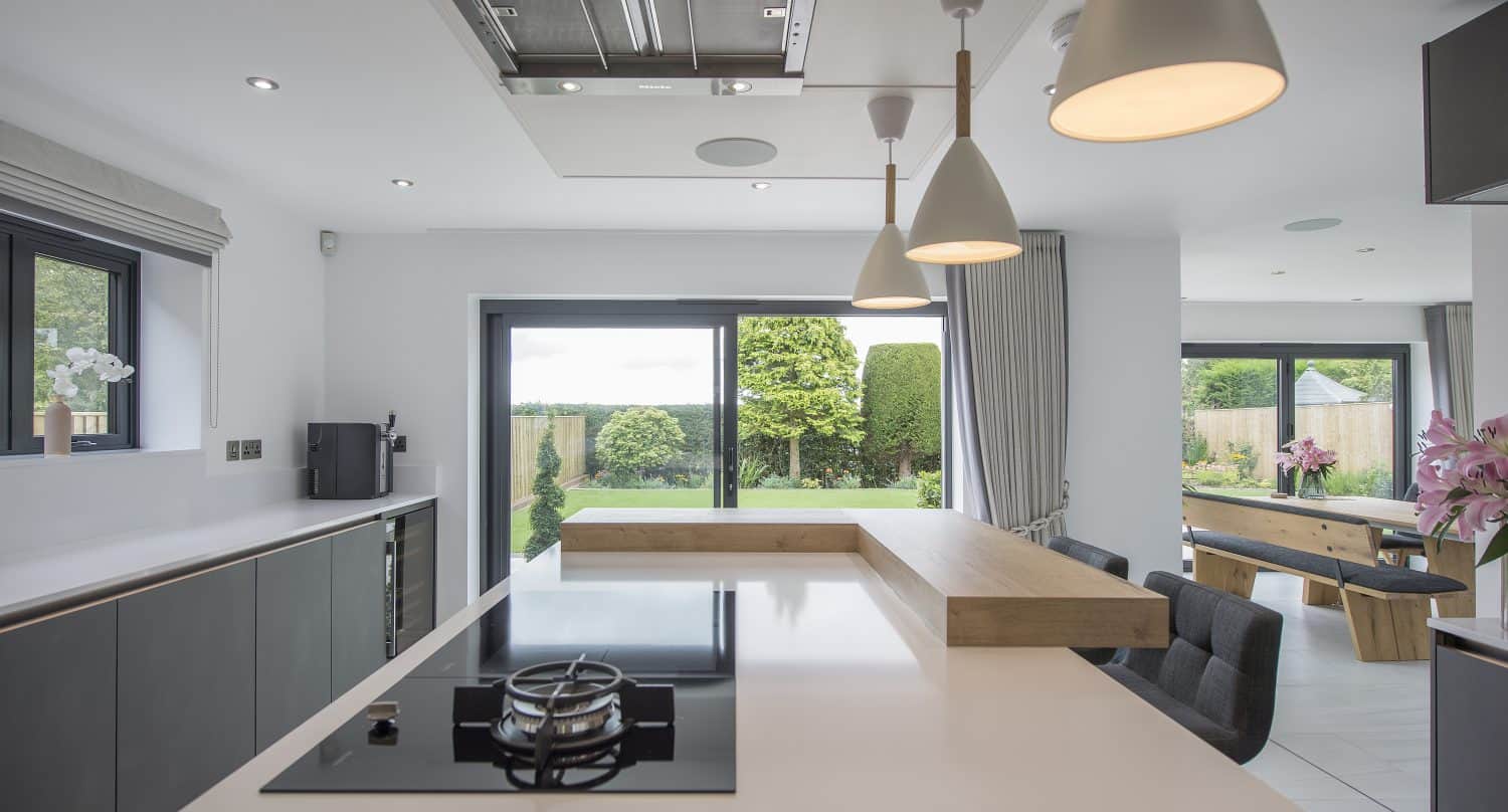 Modern kitchen with white countertops, a built-in gas stove, and hanging pendant lights. Large sliding glass doors feature bespoke glass solutions, opening to a green garden that fills the space with natural light. Dining area with dark chairs is visible in the background.