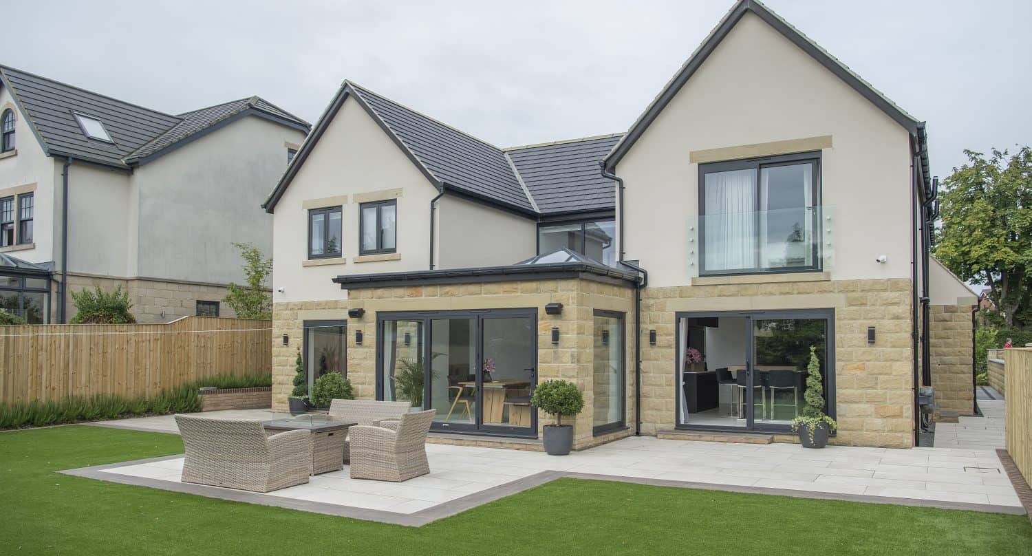 Modern two-story house with cream walls, large windows, and glass balconies, featuring sleek bifold doors opening to a patio with outdoor furniture and a well-kept artificial lawn in the backyard.
