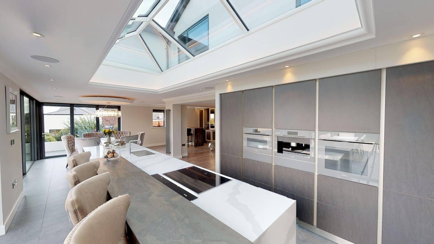 Modern kitchen with a large island, barstools, built-in appliances, and a skylight roof. Floor-to-ceiling windows and bifold doors provide natural light, while bespoke glass solutions define the dining area visible in the background.