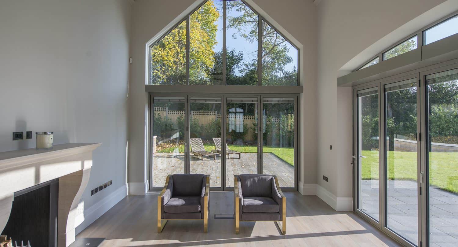 Spacious living room with high ceiling, large windows, bifold doors, two armchairs, a glass coffee table, a fireplace, and a view of a sunny garden with trees and outdoor seating.