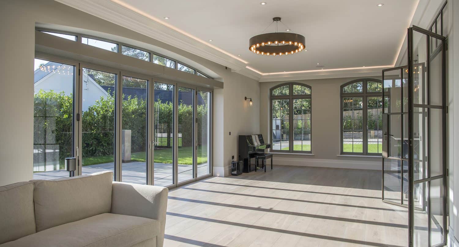 Bright, spacious living room with large windows and bespoke glass solutions, including bifold doors that let in natural light, a beige sofa, a piano, light wood floors, and views of a green garden outside.