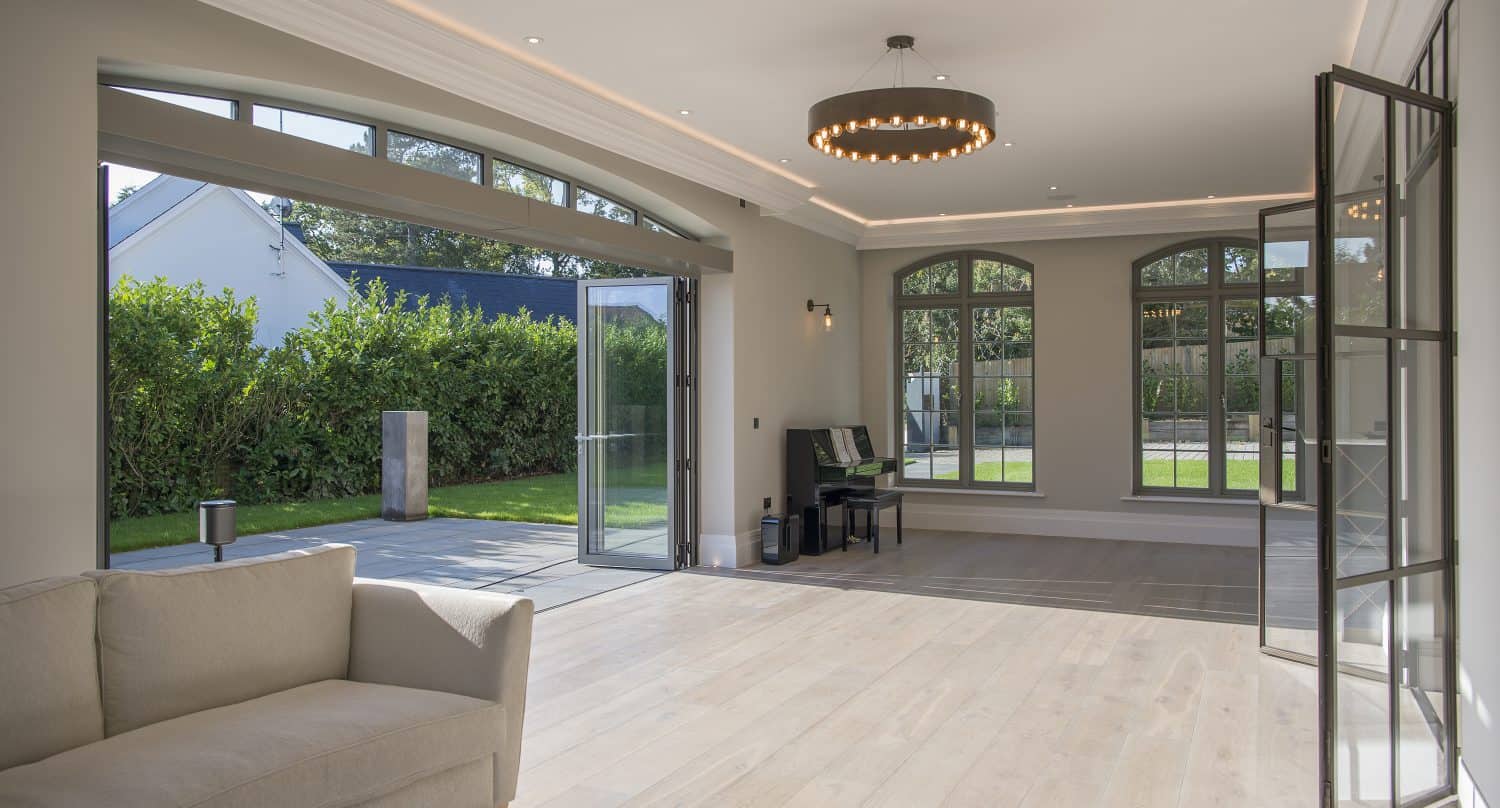 A spacious, modern living room with light wood flooring and large windows features bifold doors that open to a garden, a beige sofa on the left, and a black piano against the far wall. Natural light fills the room.