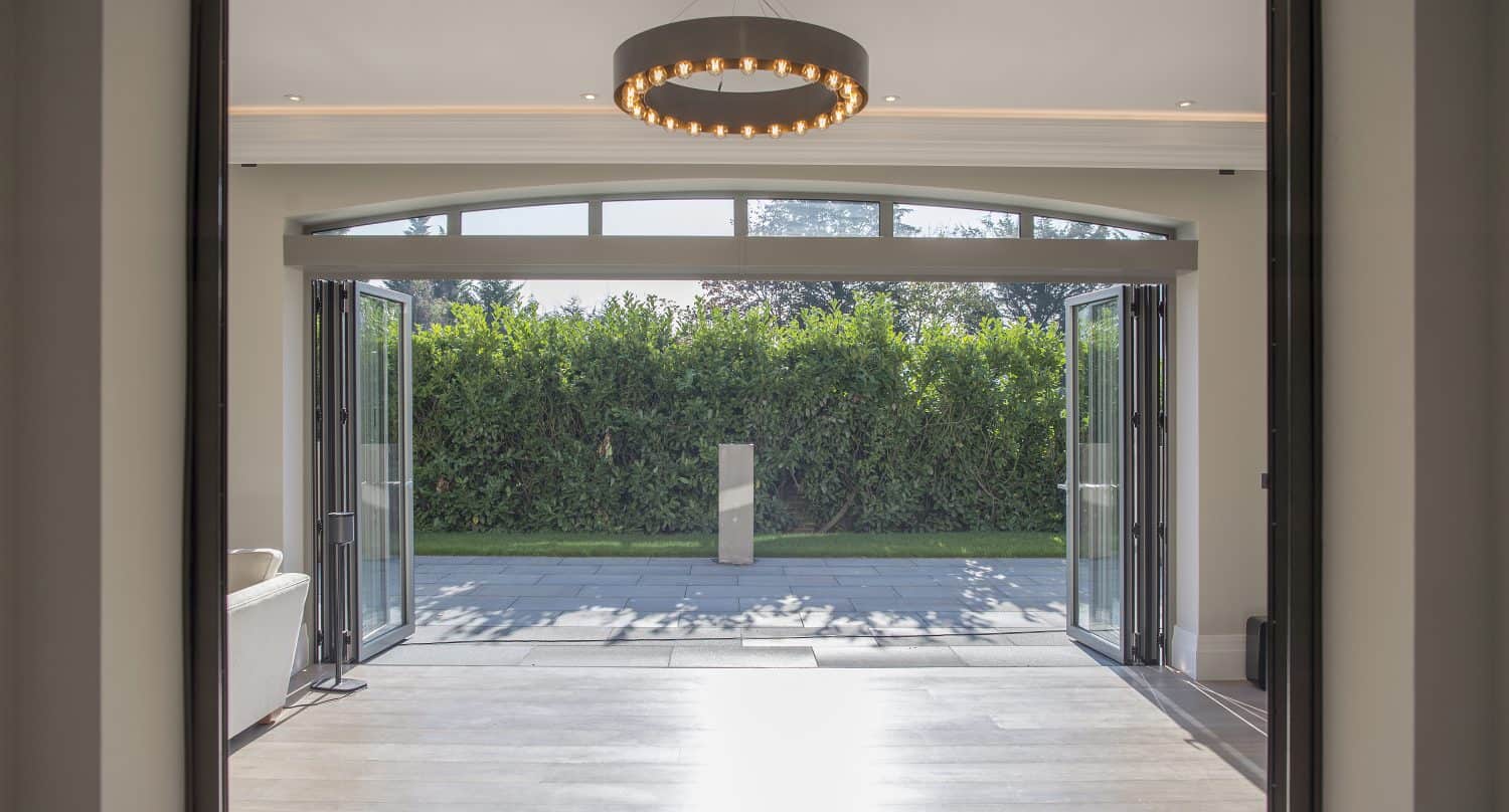 View from inside a modern room with large bifold doors leading to a patio and green hedge outside; a circular ceiling light hangs from the center, and sunlight fills the space through bespoke glass solutions.