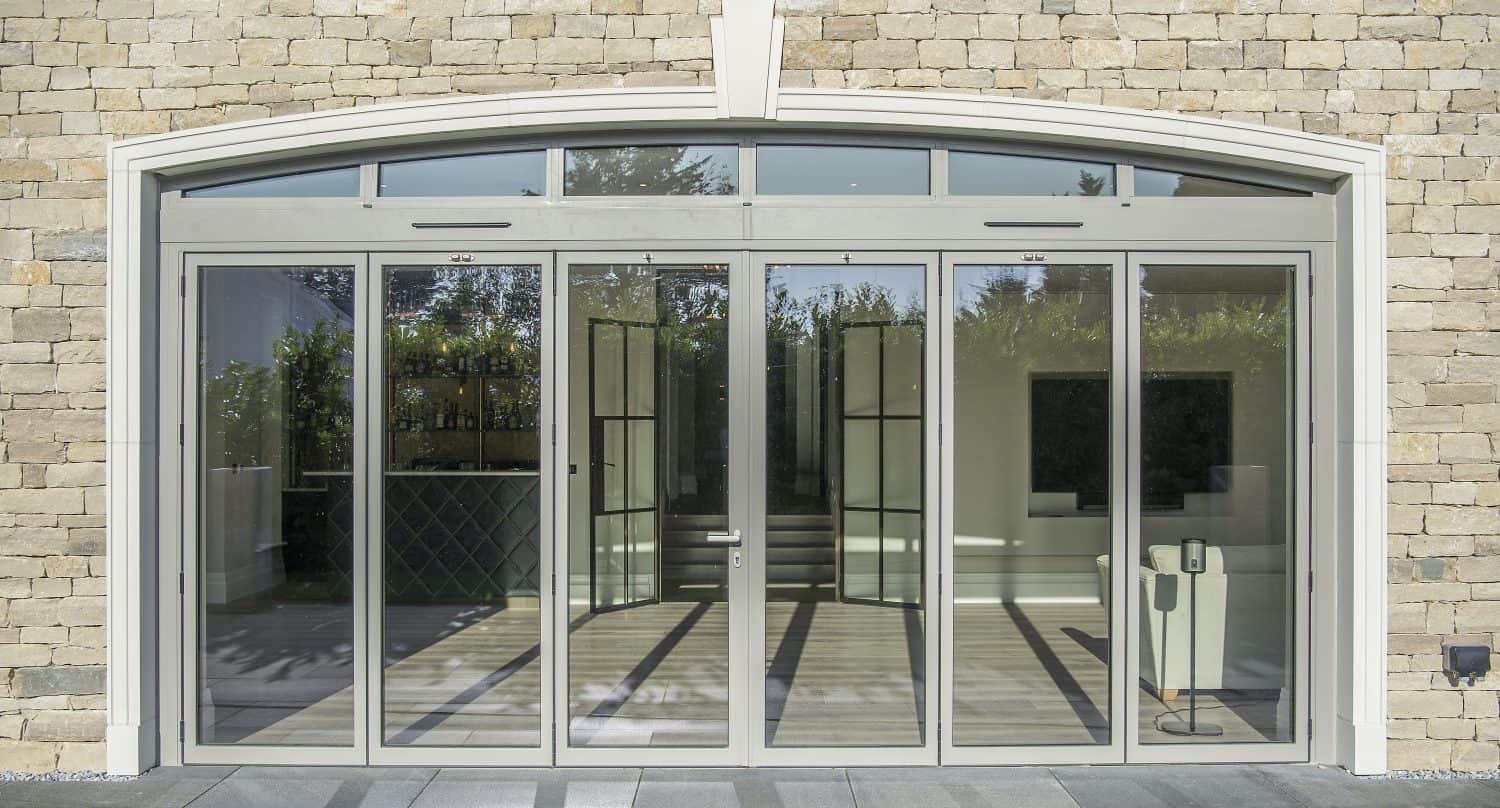 Glass double front doors with side panels and transom windows set in a stone wall, opening to a modern, minimally furnished interior with sunlight streaming in and greenery visible outside—an elegant example of bespoke glass solutions.