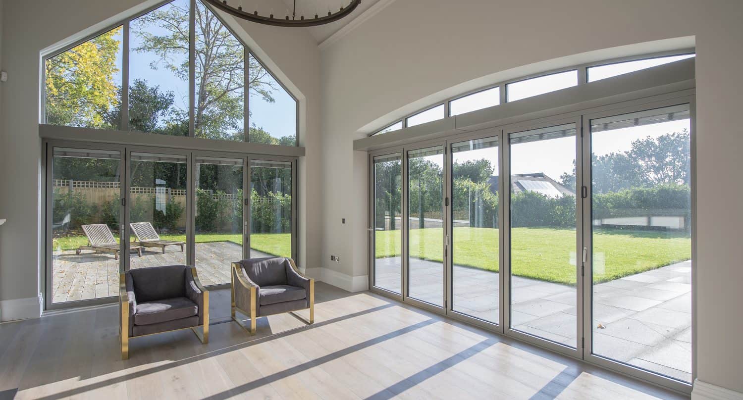 A bright, modern room with large floor-to-ceiling windows and bespoke glass solutions, plus glass doors opening to a sunny patio and green garden. Two wooden armchairs sit on light wood flooring, with sunlight streaming in.
