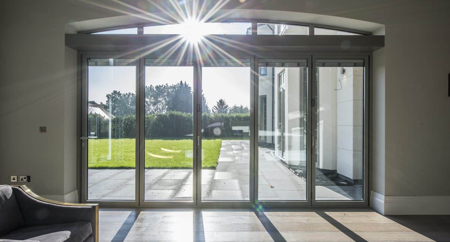 Sunlight streams through large bifold doors, illuminating a bright modern room with wooden floors and a gray armchair, and revealing a green lawn and patio outside.
