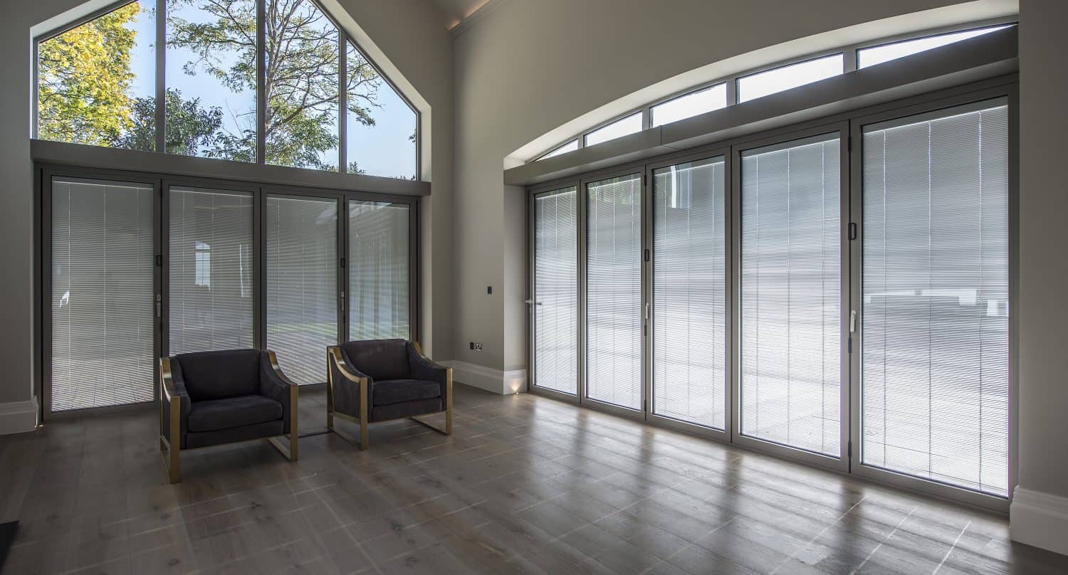 A bright, modern room with large floor-to-ceiling windows, bespoke glass solutions, two dark armchairs, light wood flooring, and a high ceiling. The view outside shows trees and blue sky.