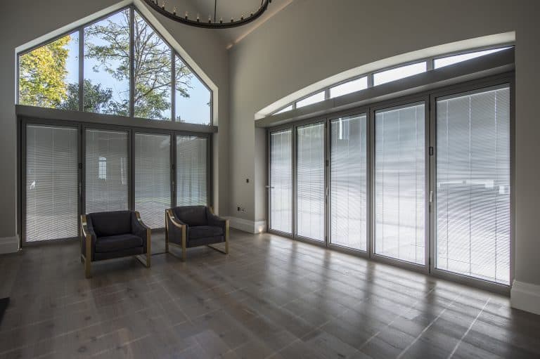 A bright, modern room with large floor-to-ceiling windows, bespoke glass solutions, two dark armchairs, light wood flooring, and a high ceiling. The view outside shows trees and blue sky.