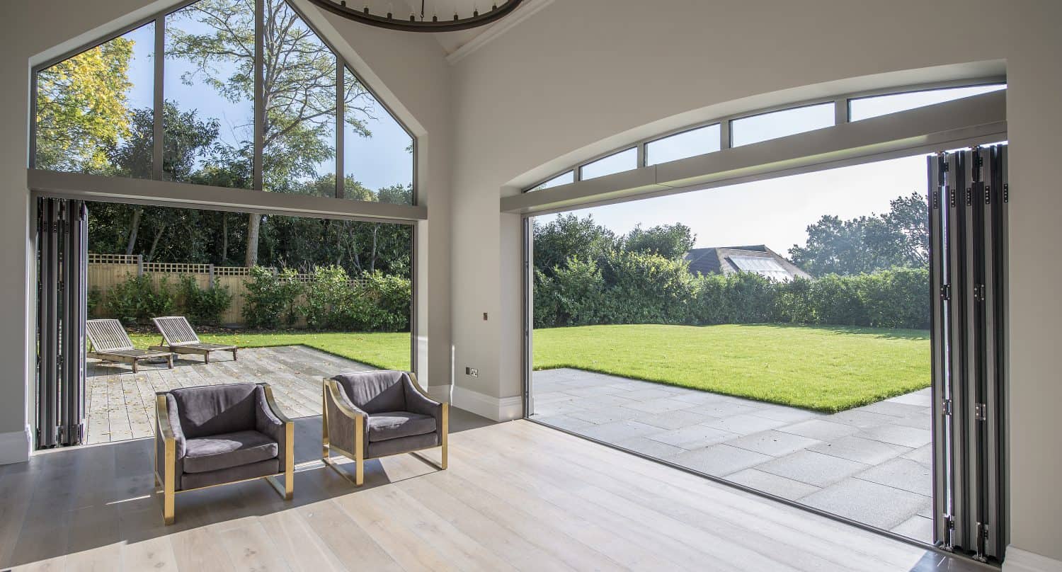 A modern, bright room with bespoke glass solutions, including large folding doors and windows opening to a sunny patio, green lawn, and trees. Two wooden armchairs are positioned by the open space, letting in natural light.