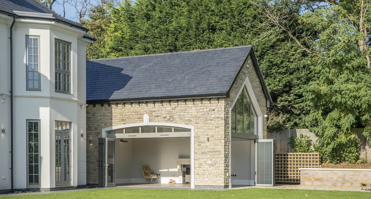Modern house extension with stone walls and large bifold doors, open to reveal a bright interior with minimal furnishings, set in a garden with green lawn and tall trees in the background.