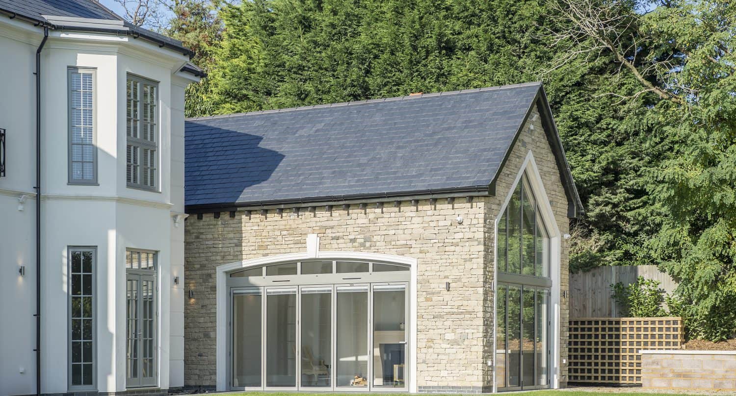 Modern house extension with large glass windows and bifold doors, stone exterior walls, and a slate roof. The extension connects to the main white house and overlooks a green lawn with trees and a wooden fence in the background.