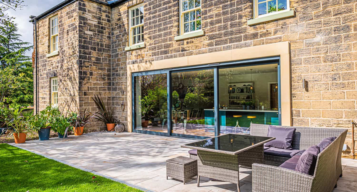 A sunlit patio with outdoor furniture sits beside a large stone house. Bifold doors open to a modern kitchen, and potted plants decorate the tiled patio area, bordered by a well-kept green lawn.