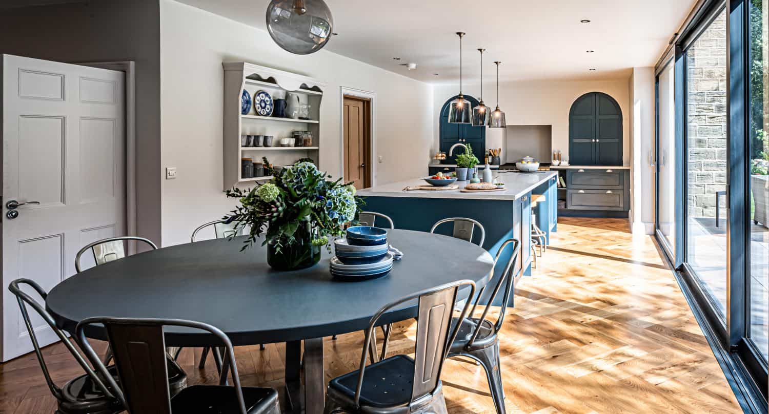 Modern kitchen with blue cabinets, a round dining table set with dishes and metal chairs, wooden herringbone floor, pendant lights, open shelves, and large sliding doors letting in natural light.