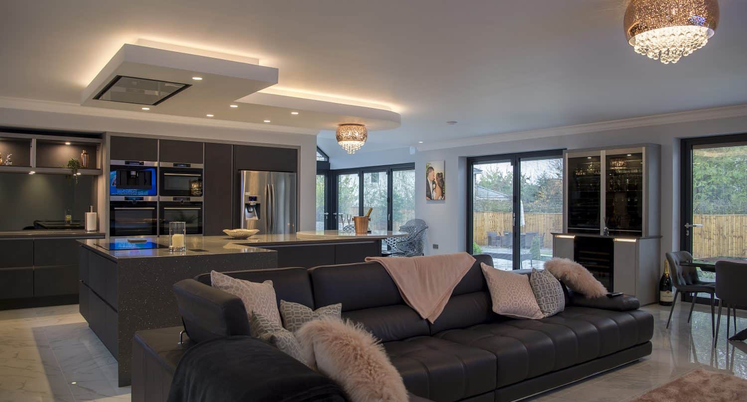 Modern open-plan kitchen and living room with a dark leather sectional sofa, plush pillows, large island, built-in appliances, and floor-to-ceiling windows featuring bespoke glass solutions overlooking a fenced backyard. Elegant ceiling lights add warmth to the space.
