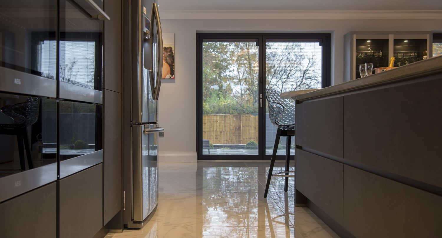 Modern kitchen with glossy marble floor, built-in appliances, a stainless steel fridge, and a high chair by sliding doors overlooking a fenced backyard with trees.