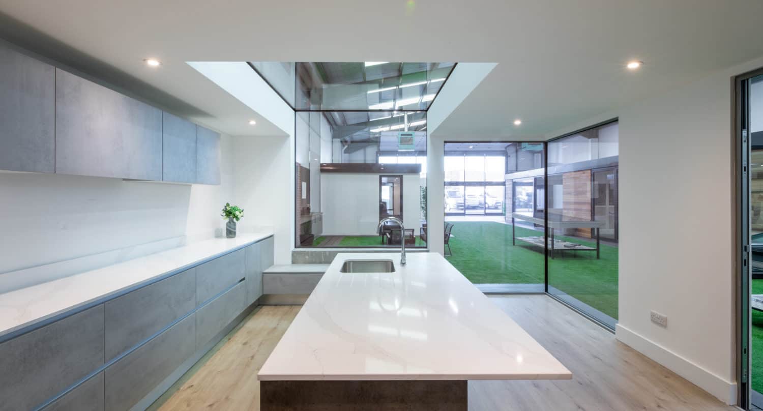 Modern kitchen with a large white island, grey cabinets, wooden floor, and a window wall overlooking a room with green artificial grass. Glass roofing and spotlights provide bright, natural lighting. Bifold doors offer seamless indoor-outdoor flow.