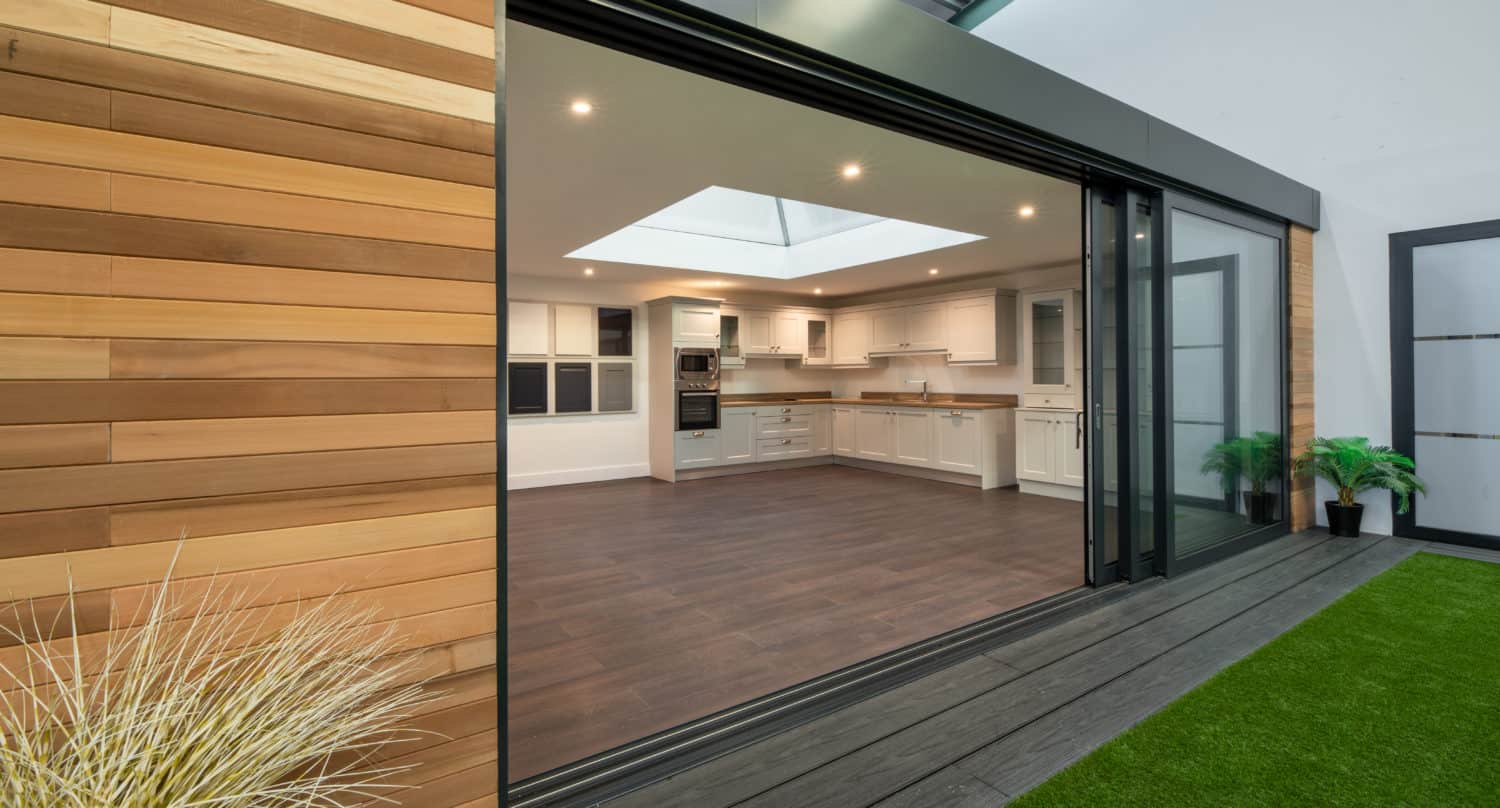 Modern open-plan kitchen with white cabinets and wooden countertops, viewed through large sliding doors. The room features a skylight, wood flooring, and opens onto a patio with artificial grass and potted plants.