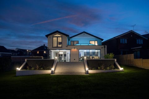 Modern two-story house with large windows, bifold doors, and outdoor lighting, viewed at dusk. The home features a wide staircase, landscaped garden beds, and is flanked by neighboring houses under a dusky blue sky.