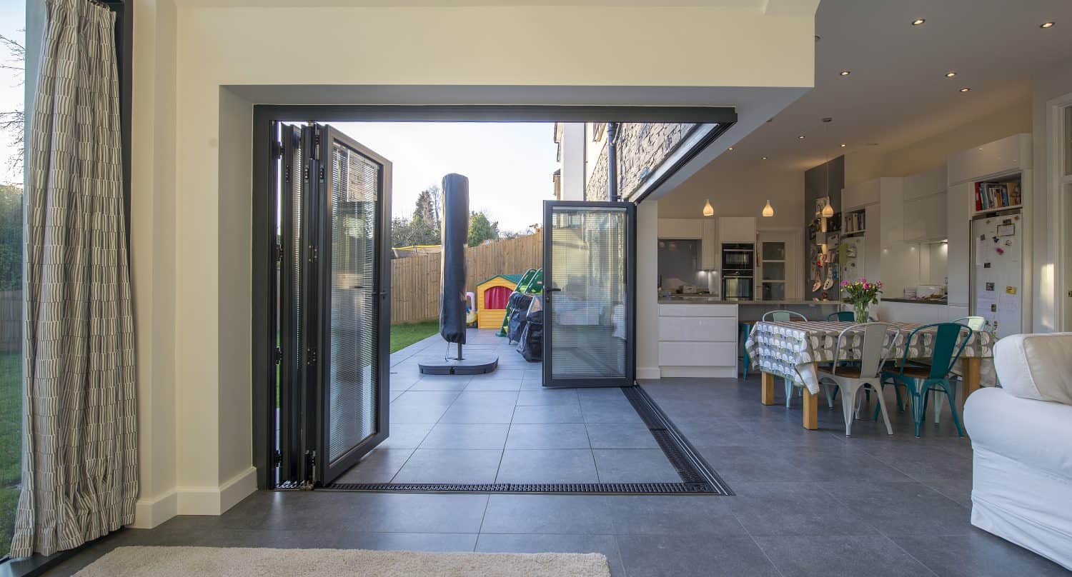 Modern open-plan kitchen and dining area with large bifold doors opening to a garden patio, blending indoor and outdoor spaces. Light-filled room with tiled floors, white cabinetry, and contemporary furnishings.