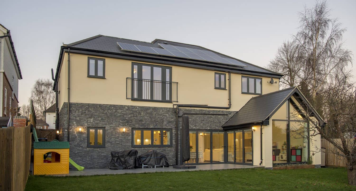 A modern two-story house with large windows and bespoke glass solutions, solar panels on the roof, and a stone accent on the lower exterior stands beside a lawn with a children’s playhouse and slide in the backyard.