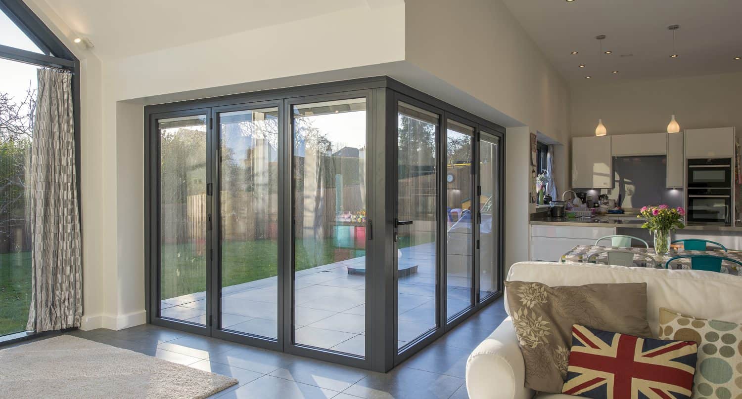 A modern living space with large bifold doors opening to a garden, a white sofa with a Union Jack pillow, a dining table with flowers, and an open-plan kitchen in the background.