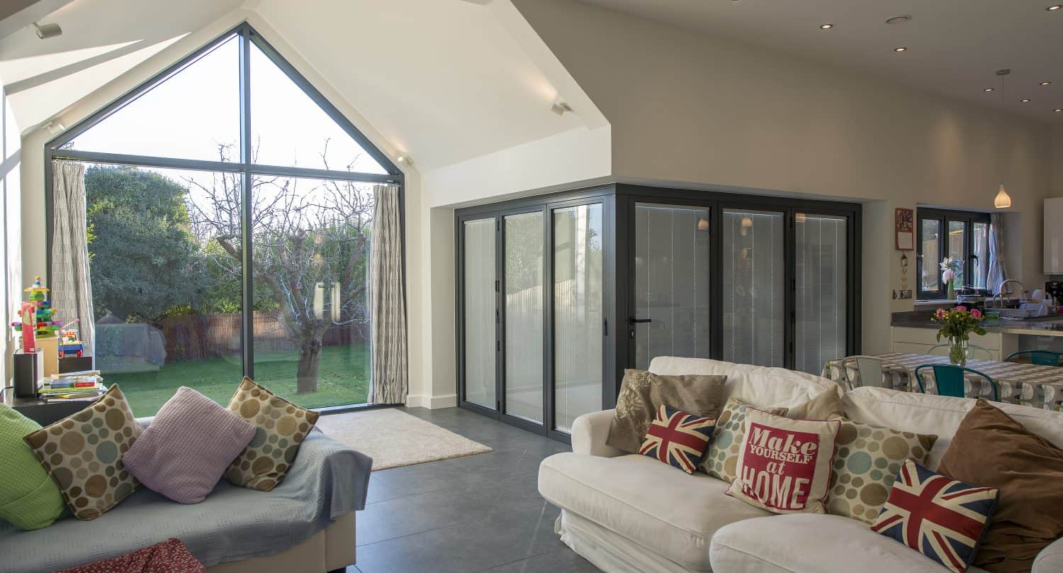 A modern living room with large floor-to-ceiling windows featuring bespoke glass solutions, white sofas with Union Jack pillows, a light blue couch, and views of a green backyard. The space is open and bright with gray tile floors.