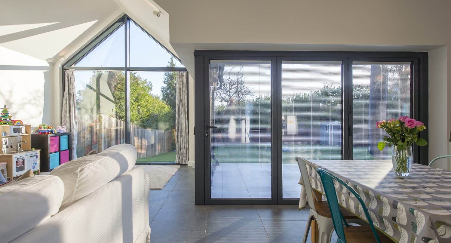 A bright living and dining area with large bifold doors and windows allows natural light to flood in. A sofa faces the glass, a table with chairs and a vase of flowers sits nearby, and children’s toys are in the corner.