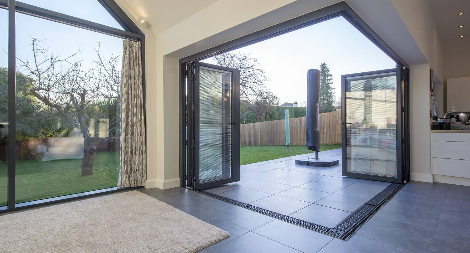 Modern home interior with large bifold doors opening to a garden. The room features tiled and carpeted floors, abundant natural light, and views of trees and a fenced yard outside, enhanced by bespoke glass solutions.